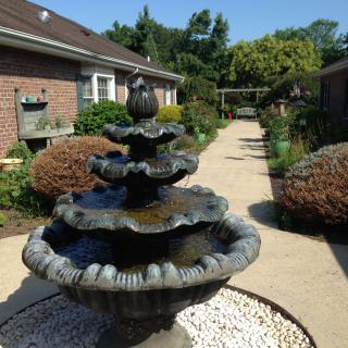 A serene garden with a tiered stone fountain and a brick building in the background.