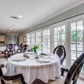 An elegantly set dining room with a round table, surrounded by chairs and illuminated by natural light from large French doors.