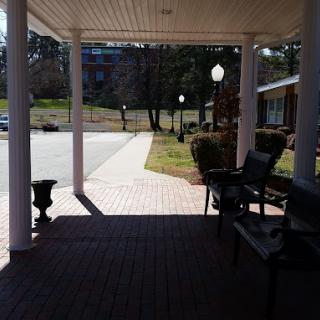 A shaded porch area with benches overlooks a sunlit pathway lined with lamp posts.