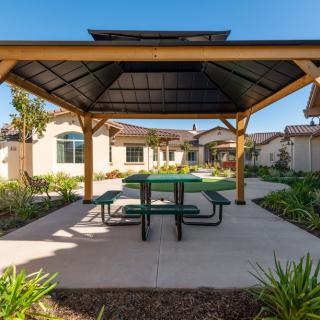 A wooden gazebo with a green picnic table underneath, surrounded by a landscaped garden on a sunny day.