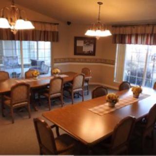 A cozy dining room with large tables and chairs, illuminated by overhead lighting and natural light from the windows.