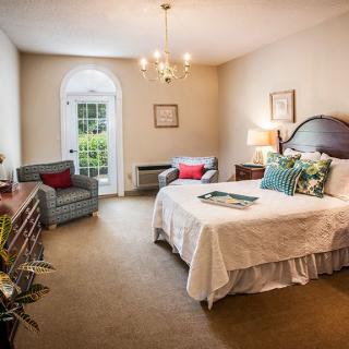A cozy and elegantly furnished bedroom featuring a bed with decorative pillows, a chandelier, and armchairs by the window.