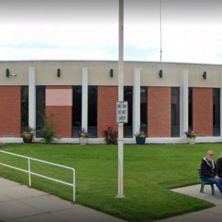 A person sitting on a bench outside a brick building with large windows and a street sign nearby.
