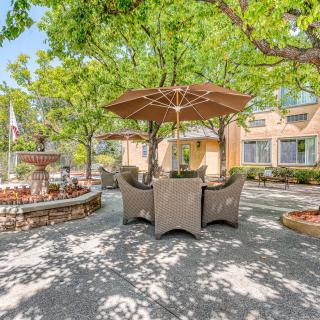 A sunny outdoor patio area with seating and a large umbrella, surrounded by trees and gardens.