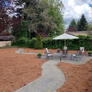 A cozy backyard patio with seating and an umbrella, surrounded by trees and mulch.