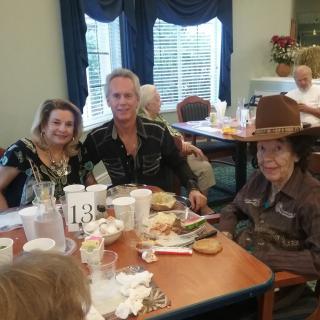 A group of people enjoying a meal together, with one wearing a large hat, in a cozy dining setting.