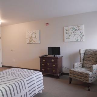 A cozy bedroom featuring a bed, patterned chair, dresser with a television, and decorative paintings on the wall.
