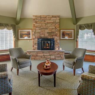 A cozy living room with a central fireplace, surrounded by comfortable armchairs and a small coffee table, illuminated by natural light through large windows.