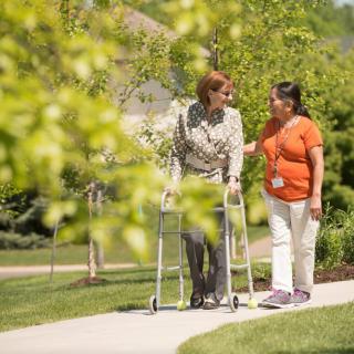 A caregiver assists an elderly person with a walker outdoors on a sunny day.
