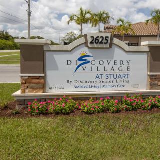 Entrance sign for Discovery Village at Stuart, featuring a logo and address with surrounding landscape.