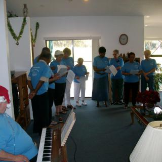 A group of people in blue shirts performing a choir indoors with a pianist accompanying them.