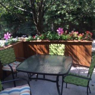 A cozy outdoor patio with a glass table and chairs, surrounded by lush plants and vibrant flowers.