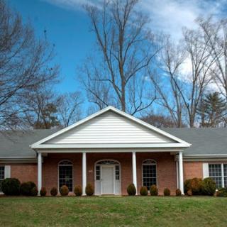 A suburban brick house with a white gabled roof and front porch, set against a backdrop of trees and blue sky.