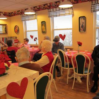 A cozy indoor setting where seniors gather for a meal, adorned with Valentine's decorations and a warm atmosphere.