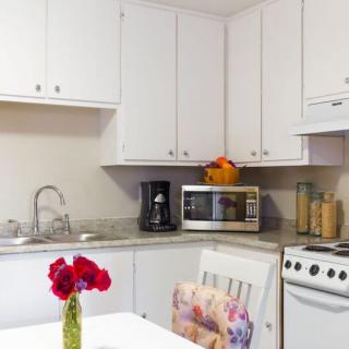A cozy kitchen with white cabinets, featuring a coffee maker, microwave, and stove. A vase of red flowers decorates the table.