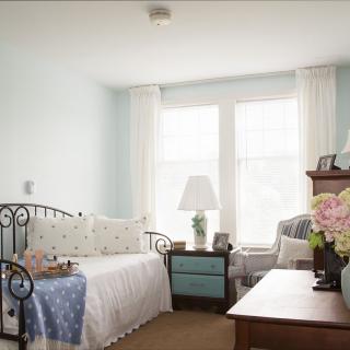 A bright and cozy bedroom featuring a daybed with decorative pillows, a side table with a lamp, and a floral arrangement on the coffee table.