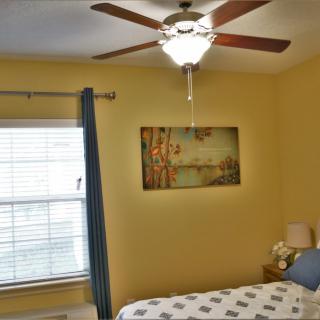 A cozy bedroom with a ceiling fan, window, curtains, and a painting above the bed.