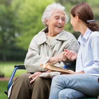 An elderly woman in a wheelchair shares a joyful conversation with a young woman in a park.