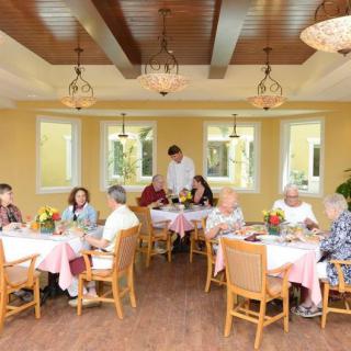 A cozy dining room where seniors are enjoying a meal, served by a waiter.