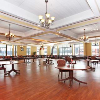 A spacious dining area with elegant chandeliers and wooden flooring, featuring several tables and chairs, surrounded by large windows.