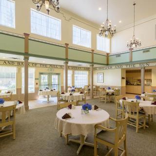 A spacious dining room with multiple round tables, chairs, and large windows allowing natural light.