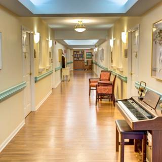 A well-lit hallway in a nursing home featuring a piano and chairs along the walls.