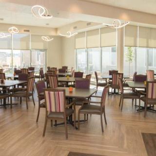 A modern dining area with tables and chairs, illuminated by natural sunlight through large windows.