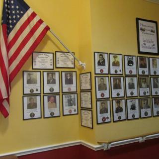 A wall display featuring an American flag, certificates, and framed photos arranged on a yellow background.