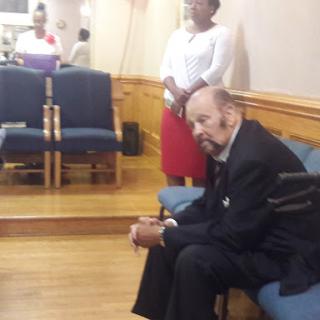 An elderly man seated in a church while a woman stands nearby.