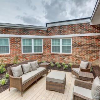 A cozy outdoor patio with wicker furniture and cushions, set against a brick wall backdrop under a cloudy sky.