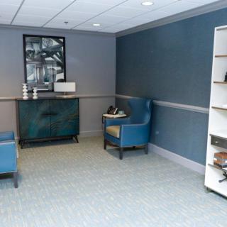 A modern waiting room with blue chairs, a shelf with books and decor, and monochrome artwork on the wall.