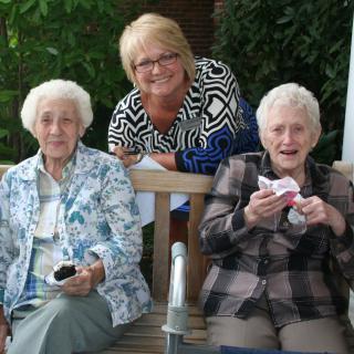 Three women enjoying an outdoor gathering, sitting on a bench and smiling.