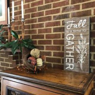 A cozy fall-themed mantel display with candles, a basket of pumpkins, and a wooden 'Fall Gather' sign against a brick wall.