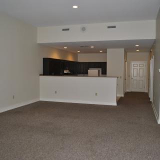 An empty apartment living room with a view of the kitchen area, featuring carpeted flooring and recessed lighting.