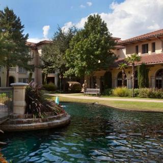 A picturesque outdoor scene featuring a building with arched windows, a tranquil pond, and lush green trees under a blue sky.