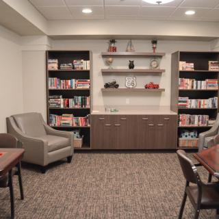 A cozy library with bookshelves, comfortable chairs, and tables in a well-lit room.