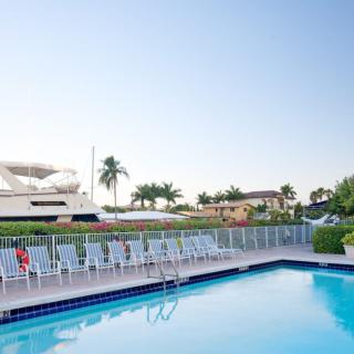 A serene outdoor pool area with lounge chairs, a yacht docked nearby, and palm trees under a clear sky.