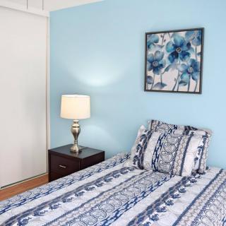 A cozy bedroom with a blue-themed decor, featuring a neatly made bed, a lamp on a nightstand, and a floral painting on the wall.