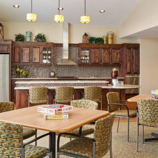 A modern kitchen interior with wooden cabinets, a countertop with bar stools, and a seating area with a table and chairs.