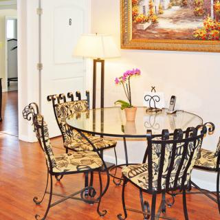 A cozy dining area with a glass-top table and ornate chairs, decorated with floral cushion seats, sits on a hardwood floor beneath elegant artwork. A lamp and an orchid plant add warmth to the space.