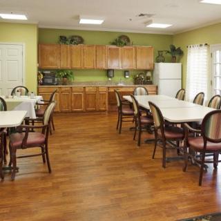 A spacious dining area with multiple tables and chairs, featuring wooden flooring and a kitchen setup with cabinets and a refrigerator.