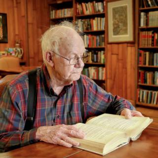 An elderly man wearing glasses and suspenders reads a book in a cozy library setting.