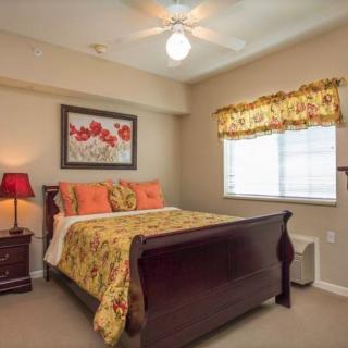 A cozy bedroom with warm tones, featuring a wooden bed, floral bedding and artwork, a nightstand with a lamp, and a window with patterned curtains.