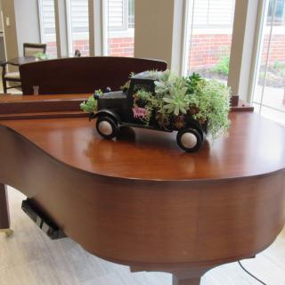 A decorative toy truck with plants sits atop a grand piano in a well-lit room.