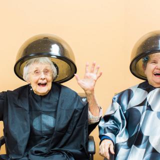 Two elderly women laughing under hair dryers at a salon.