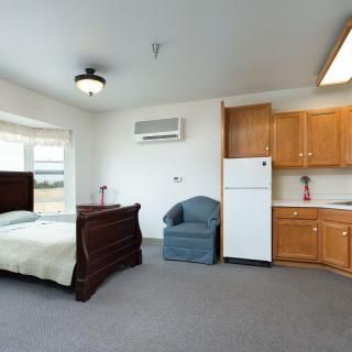 A cozy studio apartment featuring a bed, chair, and kitchenette with a refrigerator.