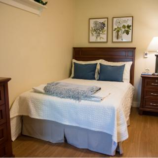A cozy bedroom featuring a neatly made bed with two blue pillows, a blanket, wooden furniture, and framed artwork on the wall.