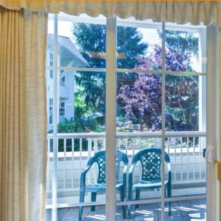 A view through sliding glass doors onto a patio with two chairs, framed by curtains and showcasing trees outside.
