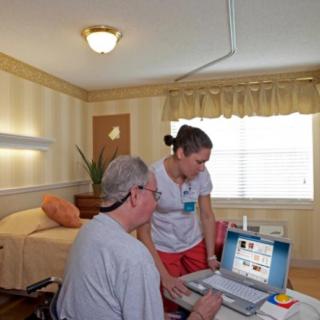A caregiver assists a patient with a laptop in a nursing home room.