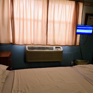 A cozy bedroom featuring a neatly made bed, a window with soft curtains, an air conditioner, and a television.